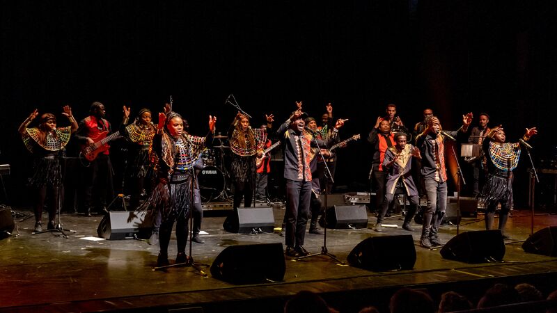 A large African music ensemble performs on a stage, with singers and instrumentalists wearing vibrant traditional clothing featuring colourful neck collars and beadwork