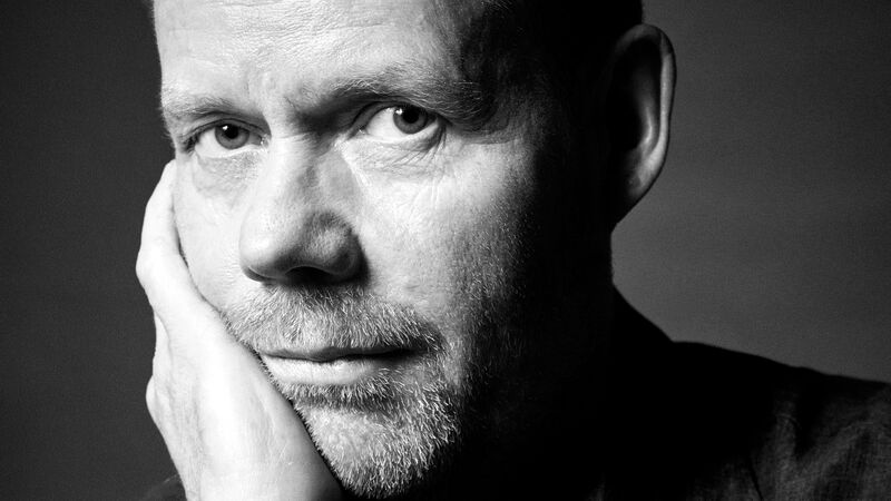 A black and white portrait of a man resting his head on his hand, looking directly at the camera with a serious expression.
