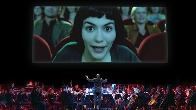 A stage showing a symphony orchestra playing below a large screen displaying a close-up of a woman from a film, looking intently at the audience