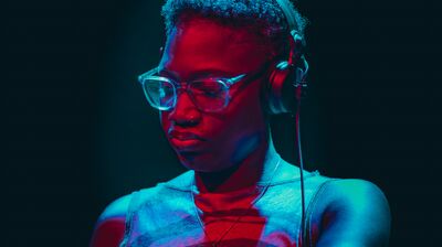 A DJ with short hair and glasses, wearing headphones, mixing under dramatic red and blue stage lighting.