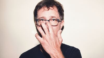 Ben Folds wearing glasses and a dark t-shirt standing in front of a white backdrop.