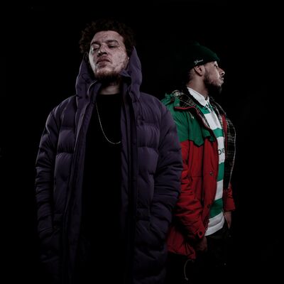 Two men pose against a black background. One wears a purple puffer jacket and the other a red and green jacket over a striped rugby shirt and beanie.