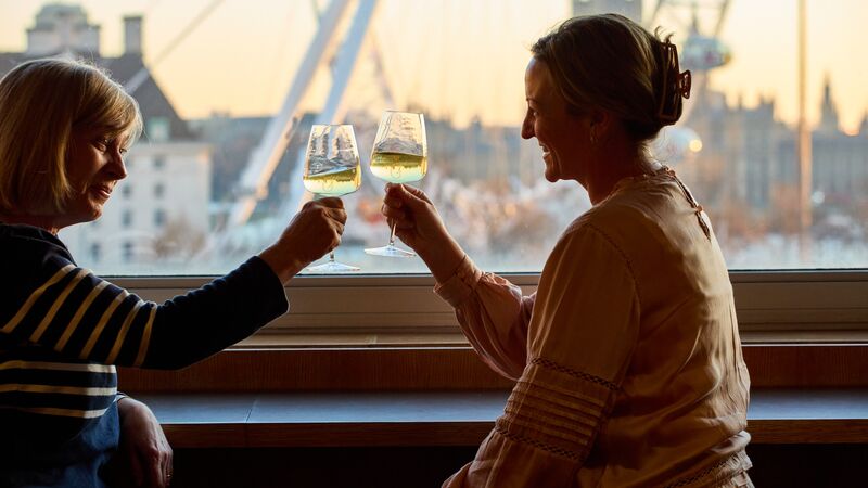 Two people seated by a window with a view of the London Eye clinking wine glasses together whilst smiling.