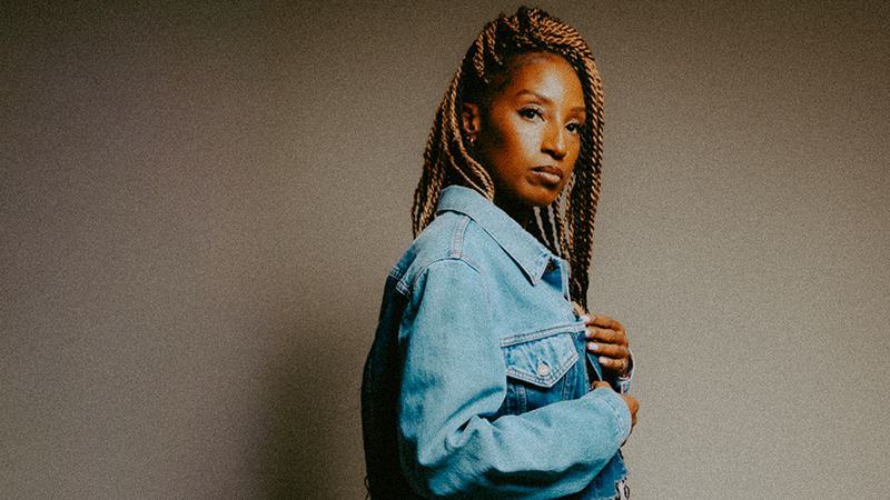A woman with long blonde braids and a denim-on-denim outfit stands side-profile, looking intently at the camera.