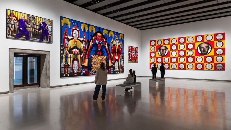 Five figures looking at artworks depicting two mens faces hung on the walls in a gallery space,