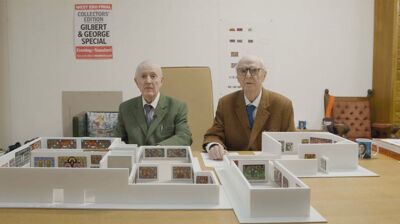 Artists Gilbert & George, two elderly white men; each wearing smart suits, sit behind a desk on top of which is a maquette of the Hayward Gallery's layout.