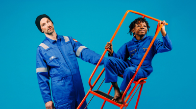 Two performers dressed in blue boiler suits stand in space together; a man holding an orange ladder which a small boy is sat in.