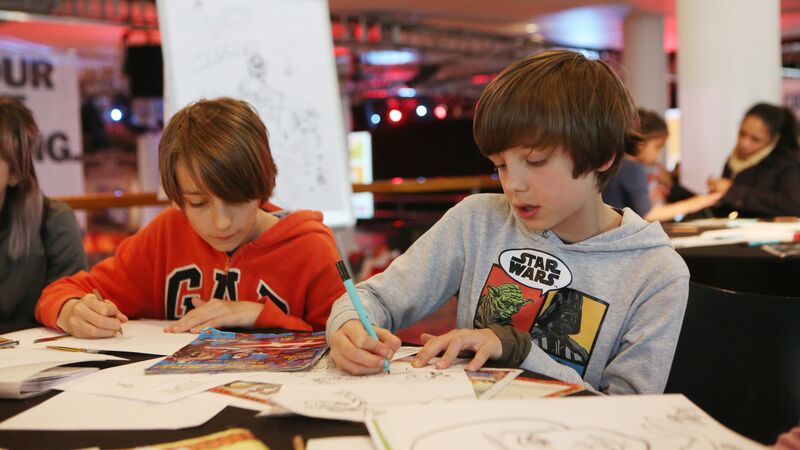 Two boys with brown hair are focused on drawing at a table. The boy in front wears a Star Wars hoodie and is coloring a picture with a blue pen.