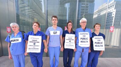 Six doctors standing united in front of JP Morgan, dressed in scrubs with paper signs reading 'Protesting for Public Health' attached to their fronts.
