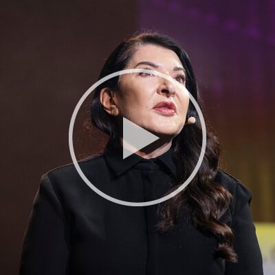Marina Abramovic, an older white woman with long dark hair over brought forward over her left shoulder