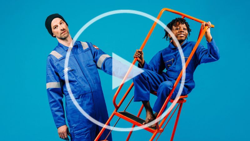 Two performers dressed in blue boiler suits stand in space together. The man holds an orange ladder which a boy is sitting on.