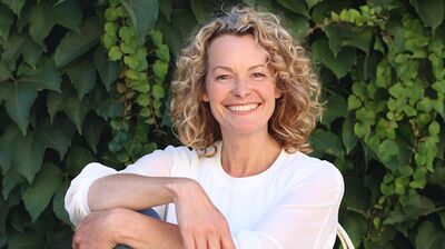 Kate Humble smiling, wearing a white top, sitting in front of green leaves