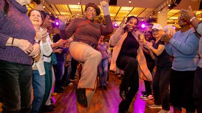 Two people dancing with one knee raised and arms lifted moving through a crowd of people clapping