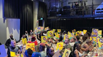 A crowded room of adults surrounded by painting materials. Many are holding up canvas with portraits they have painted on them.