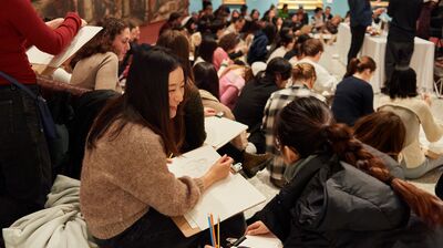 A room of people sitting, drawing. They are holding clipboards and pencils. One person looks at their neighbour and smiles.