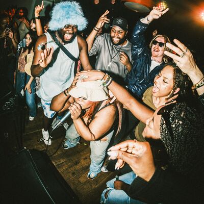 A female performer wearing a bucket hat sings into a microphone amidst a dense, joyful crowd at a lively concert or party, with a man in a light blue furry hat dancing behind her.