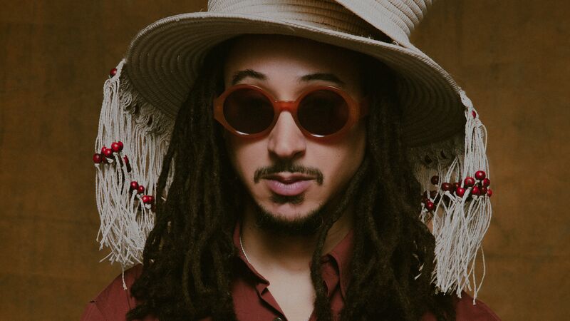 Portrait of a man with dreadlocks, wearing two stacked straw hats with tassels and beads, brown sunglasses, a terracotta shirt, and large rings.