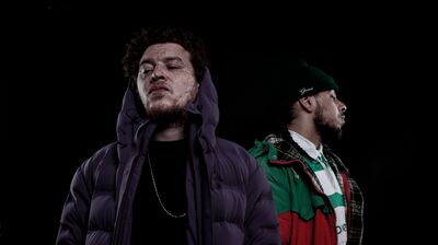 Two men pose against a black background. One wears a purple puffer jacket and the other a red and green jacket over a striped rugby shirt and beanie.