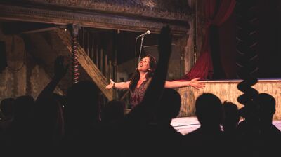 A female singer with long dark hair performs on a dimly lit, vintage stage with her arms wide open, singing passionately to a silhouetted audience.