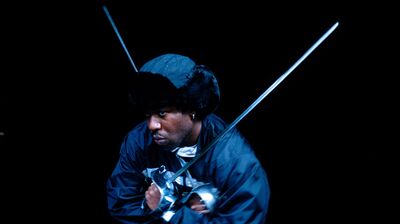A Black man wearing a fur-lined aviator hat and a black jacket wields two fencing foils against a dark background.