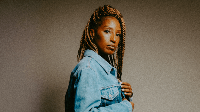 A woman with long blonde braids and a denim-on-denim outfit stands side-profile, looking intently at the camera.