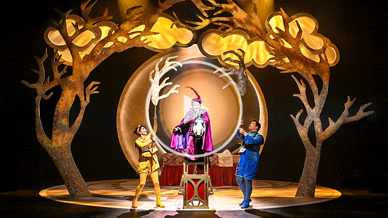 A performer wears a purple cloak as they ride a puppet horse, two other performers hold tree branches above them