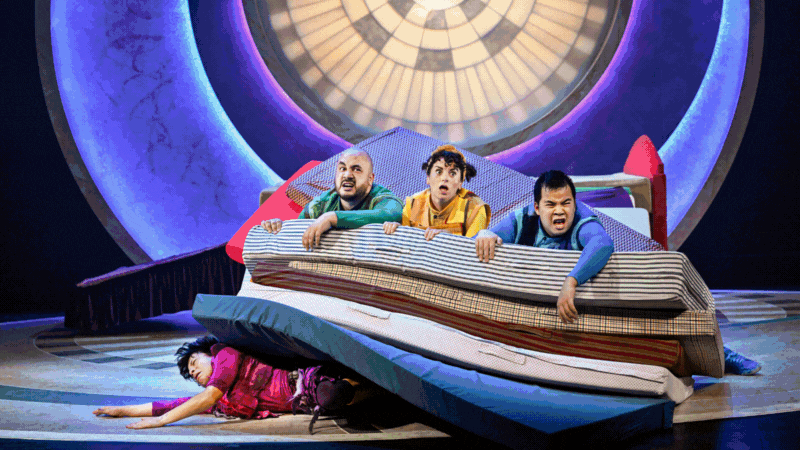 Four brightly costumed acrobats or actors, in a human chain, strain dramatically against a low red table and a backdrop of large, colorful glowing arches on a stage.