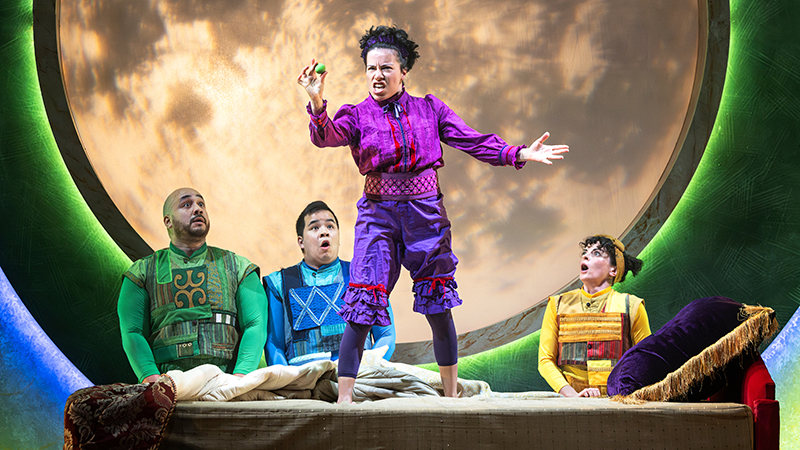 A performer in purple stands on a bed in front of a moon looking intently at a pea as three other poeple look on in surprise
