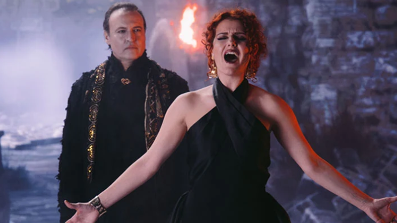 A singer with red hair caught mid-note. In the background, a man dressed in fantasy attire watches her