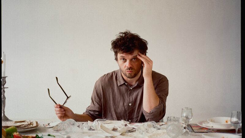 Adam Kammerling sitting at a table with his hand touching his head, holding his glasses in the other with smashed plates on the table