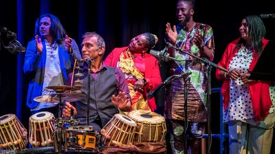 Image of five people onstage with one seated at a drum set adn the others are clapping standing behind them
