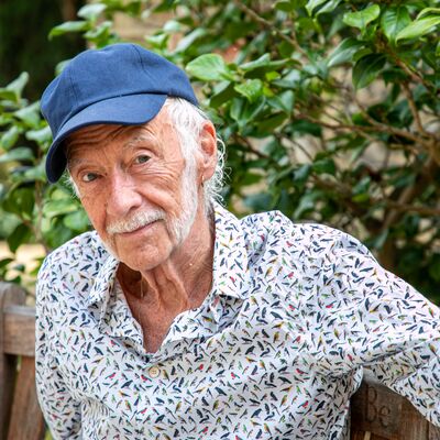 Roger McGough seated on a wooden bench wearing a blue cap