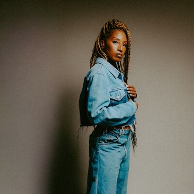 A woman with long blonde braids and a denim-on-denim outfit stands side-profile, looking intently at the camera.
