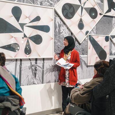 A guide in a red cardigan and hijab speaks to visitors in front of abstract geometric artwork.