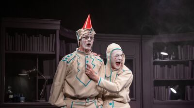 Two performers dressed in cream and blue clown outfits, with white face paint, one is pulling the elastic of a party hat the other is wearing