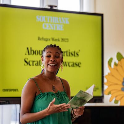Charlotte, a Black women artist smiles as she reads her own poetry in front of a yellow screen.