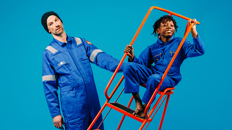 Two performers dressed in blue boiler suits stand in space together; a man holding an orange ladder which a small boy is sat in.