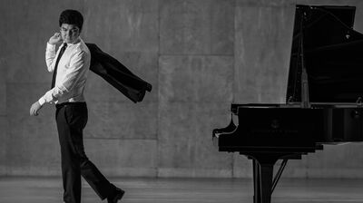 Black and white image of a man wearing a suit walking away from a grand piano with jacket over one shoulder