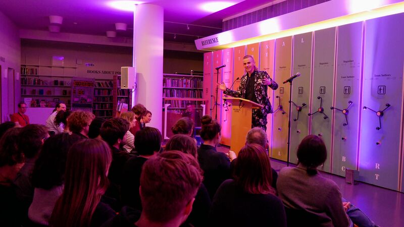 Speaker stood at a yellow podium performing in the National Poetry Library to an audience in front of colourful lockers