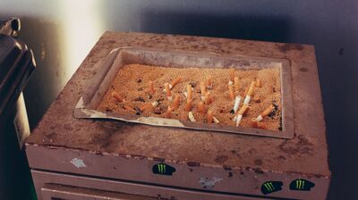 A top-down view of a rectangular sand-filled ashtray containing several cigarette butts on a rusted metal stand.