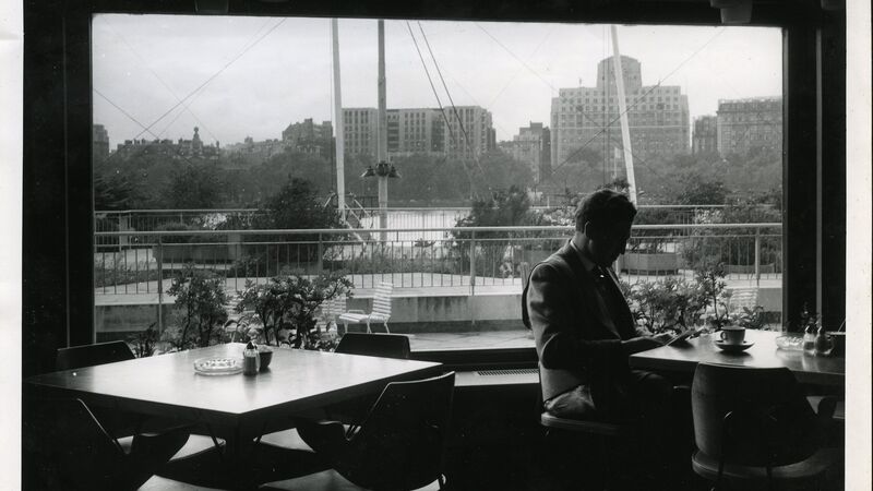 Vintage B&W photo of a man reading near a large window overlooking a city skyline and outdoor terrace.