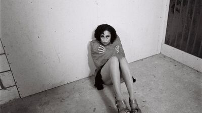 Black and white photo of a woman with curly hair sitting on a concrete floor in a bare corner, hugging herself. She is wearing fishnet tights and high-heeled platform shoes.