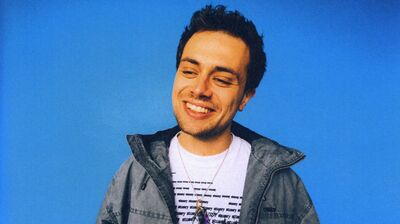 A young man with dark, wavy hair smiles widely while wearing a gray jacket and a white t-shirt with a dark graphic print and layered necklaces, against a bright blue background.
