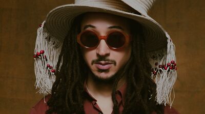 Portrait of a man with dreadlocks, wearing two stacked straw hats with tassels and beads, brown sunglasses, a terracotta shirt, and large rings.