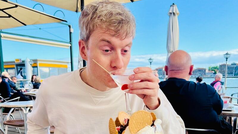 A blond man sips a martini behind a large ice cream sundae at an outdoor cafe overlooking a sunny waterway.