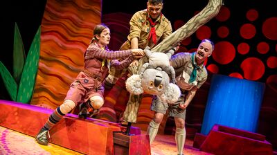 Three actors in scout uniforms help a large koala puppet on a colorful, stylized stage during a play.