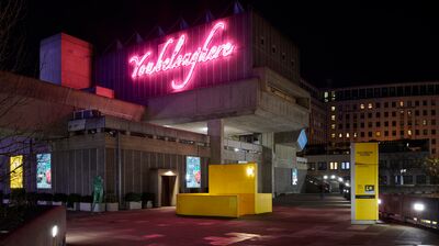 A brutalist building at night, featuring a large pink neon sign that reads "You belong here" above a yellow structure.