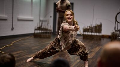 A barefoot female dancer in a brown print costume performs an aggressive lunge while holding a piece of string or rope, with a male dancer in the blurred background.
