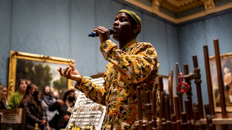 Love Ssega, wearing a multi-coloured outfit and holding a hand-held microphone as he performs inside the National Gallery; pictures with ornate frames are on the walls behind him.