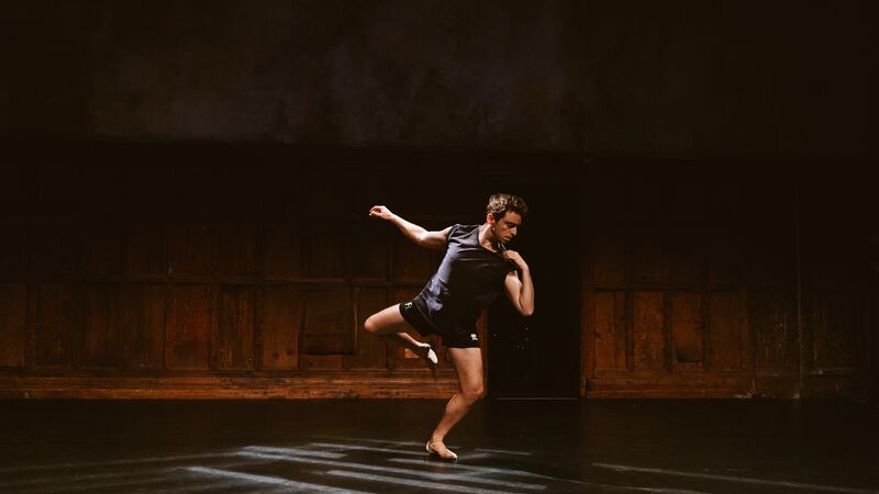 Dancer Daniel Hay-Gordon performs; he is mid-dance with one arm and one leg raised on an otherwise empty stage